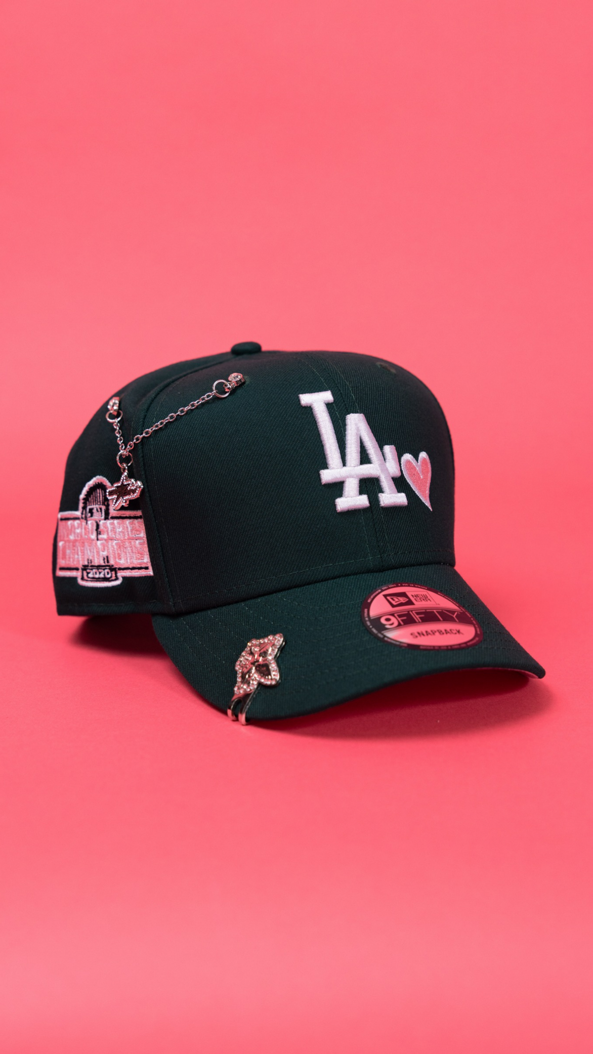 A black baseball cap with white embroidered 'LA' lettering and a pink heart, decorated with pink and gold charms, displayed on a solid pink background.